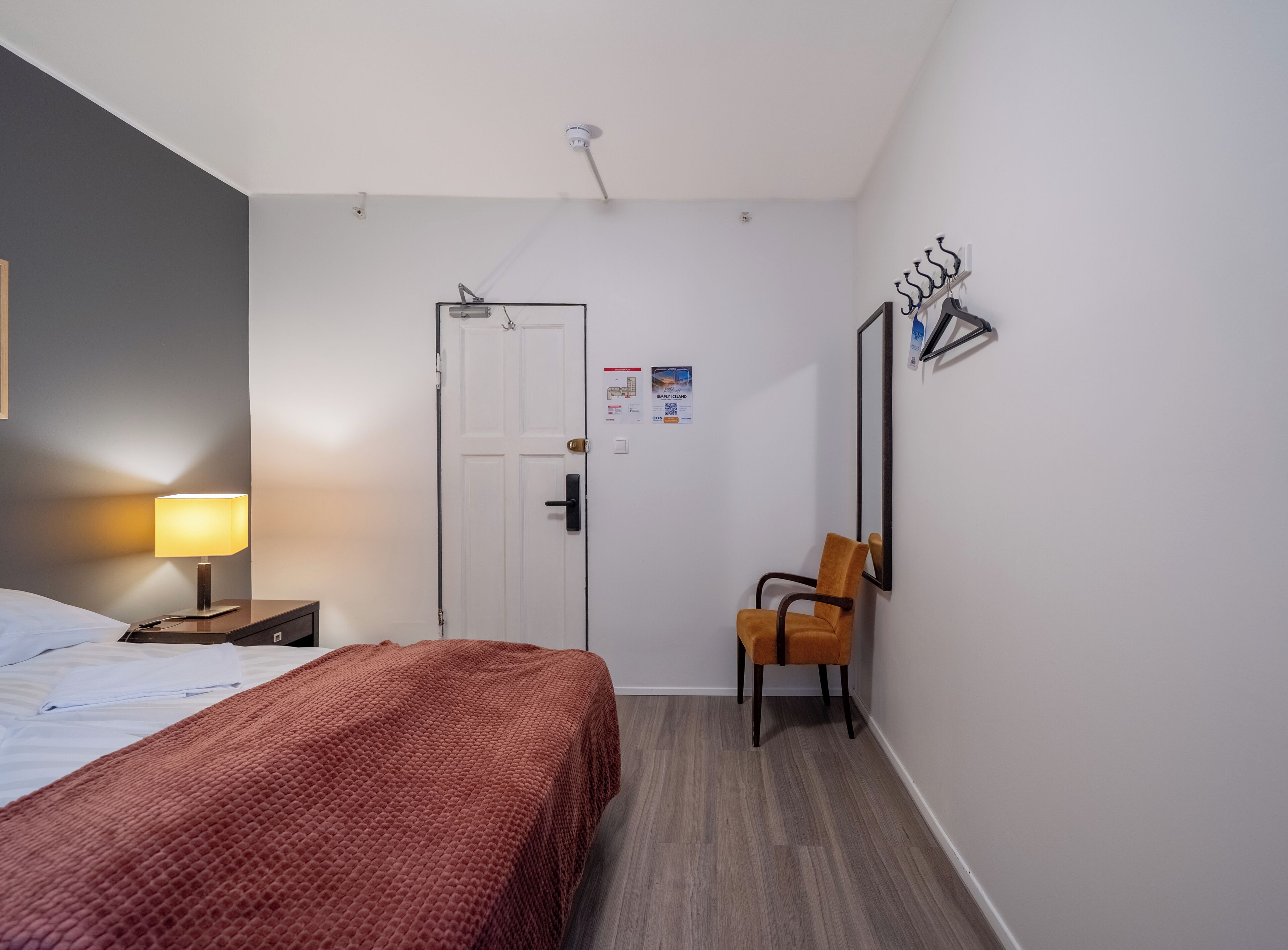 Una habitación de hotel con una cama, una mesita de noche con lámpara, una silla, una puerta y un espejo montado en la pared.
