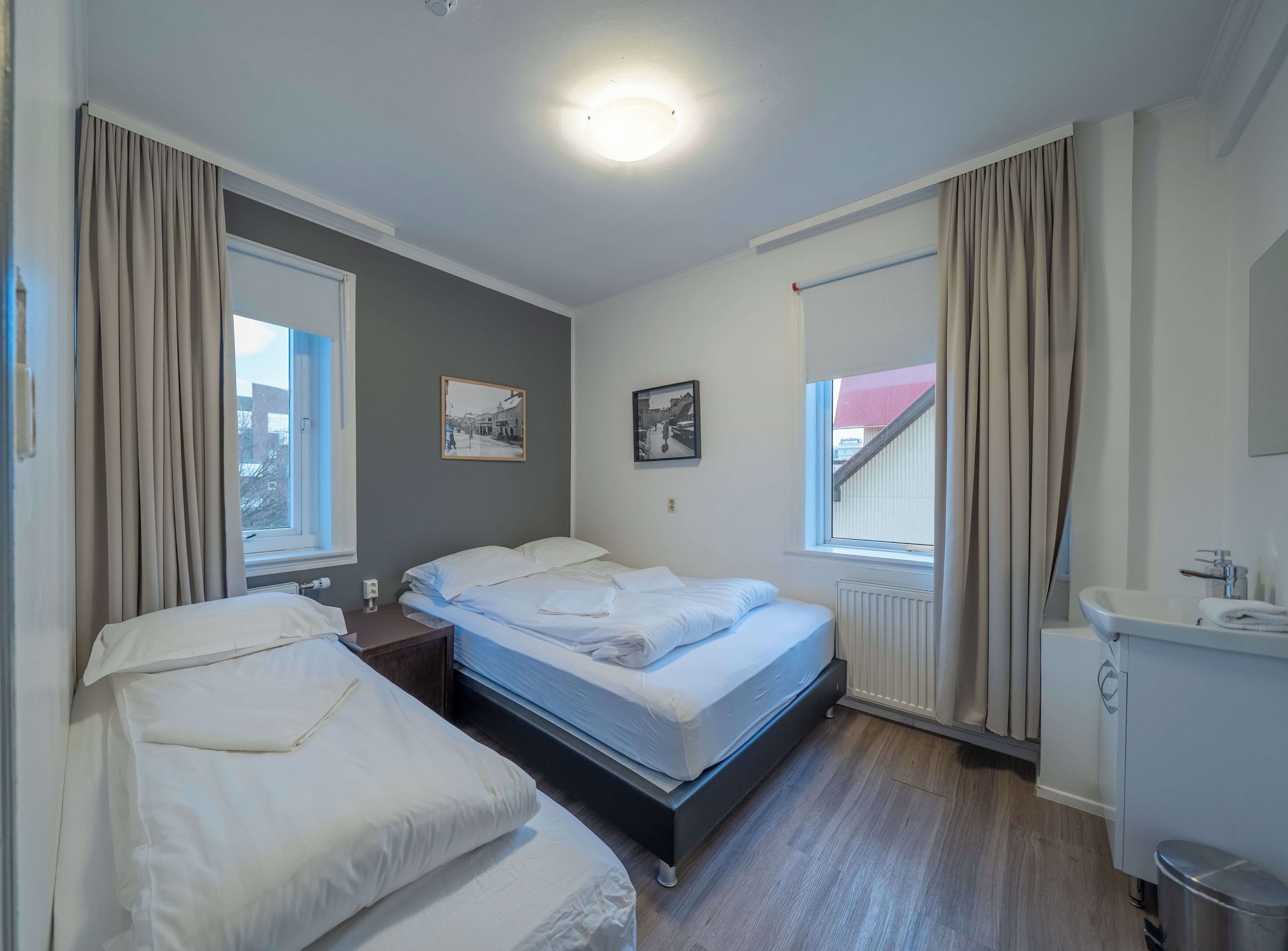 Une chambre d’hôtel avec deux lits, une fenêtre avec des rideaux, un radiateur et une petite salle de bain équipée d’un lavabo et d’un miroir.