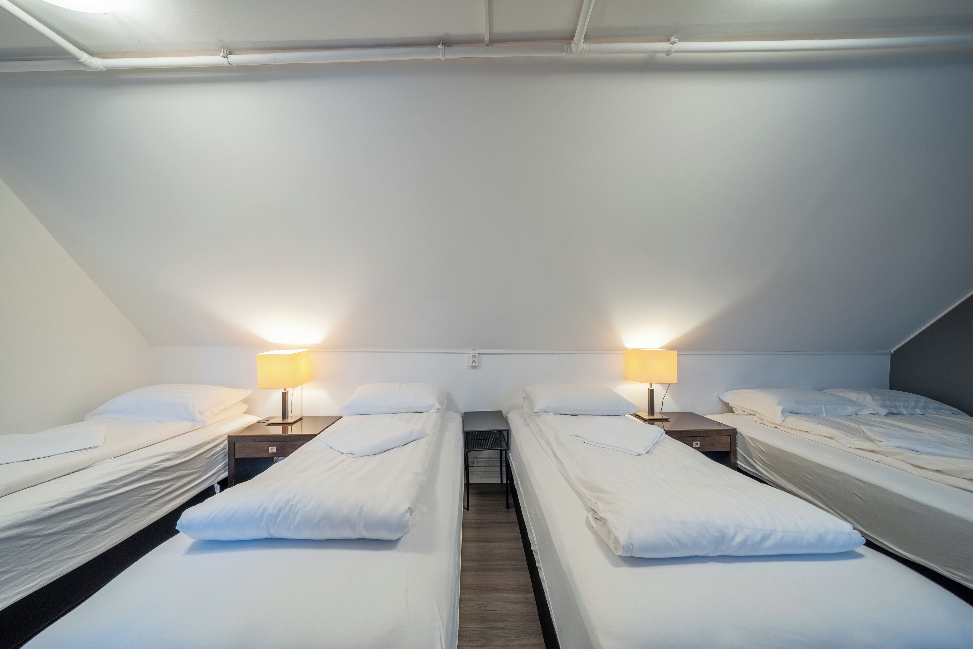 A room with four single beds arranged in a row, each with a bedside table and lamp.
