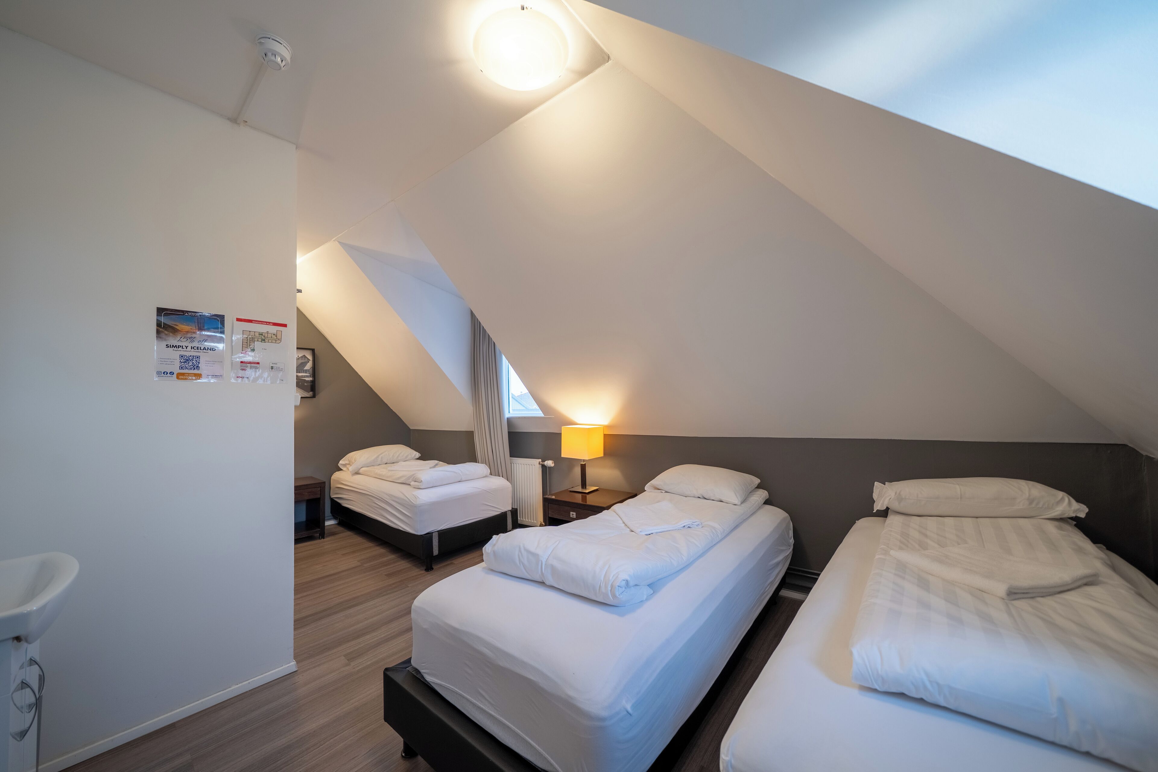 Una habitación de hotel pequeña y moderna con dos camas individuales, una mesita de noche con lámpara, un radiador y un cartel en la pared.