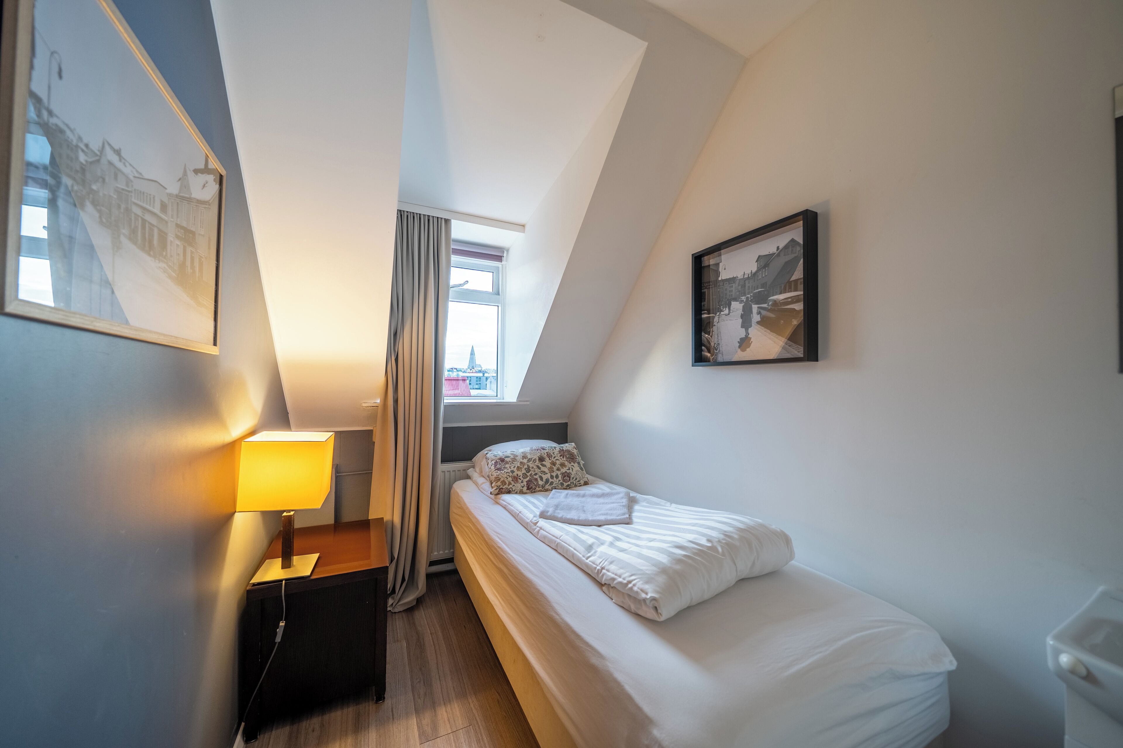A small, neatly arranged bedroom with a single bed, a bedside table, a lamp, and a window with curtains.