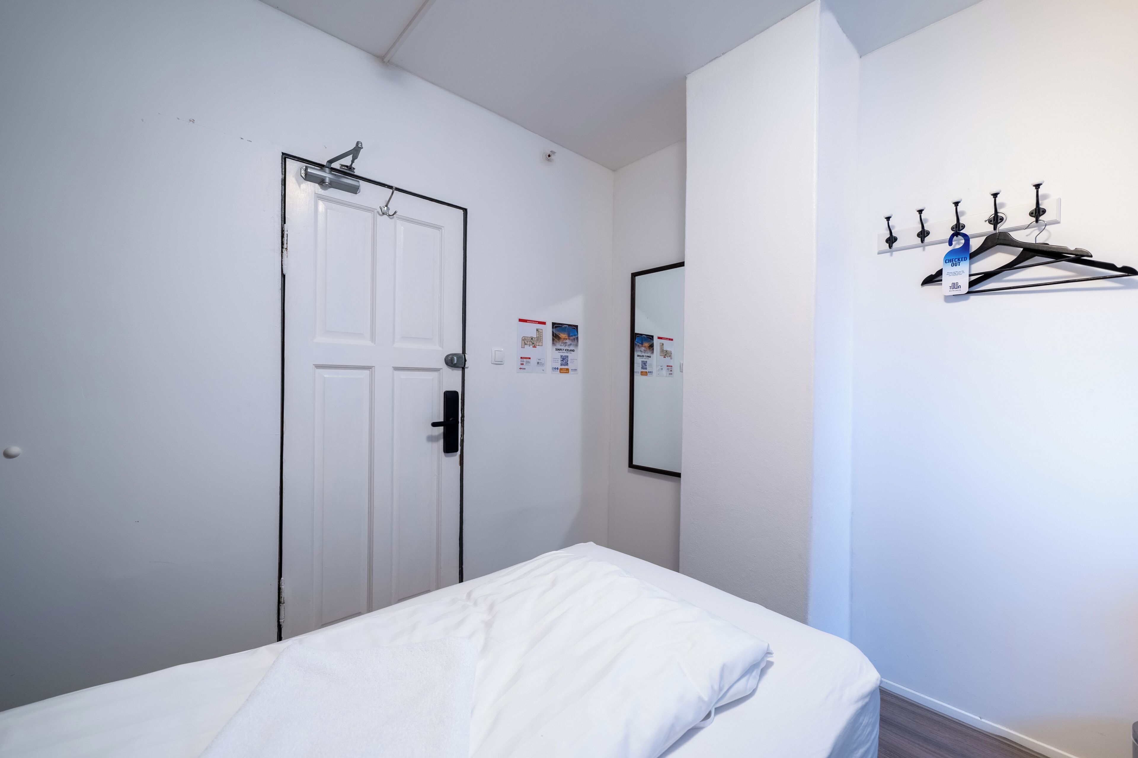 Une chambre d’hôtel avec une porte blanche, un lit avec du linge de lit blanc, un miroir au mur et un porte-manteau avec des cintres.