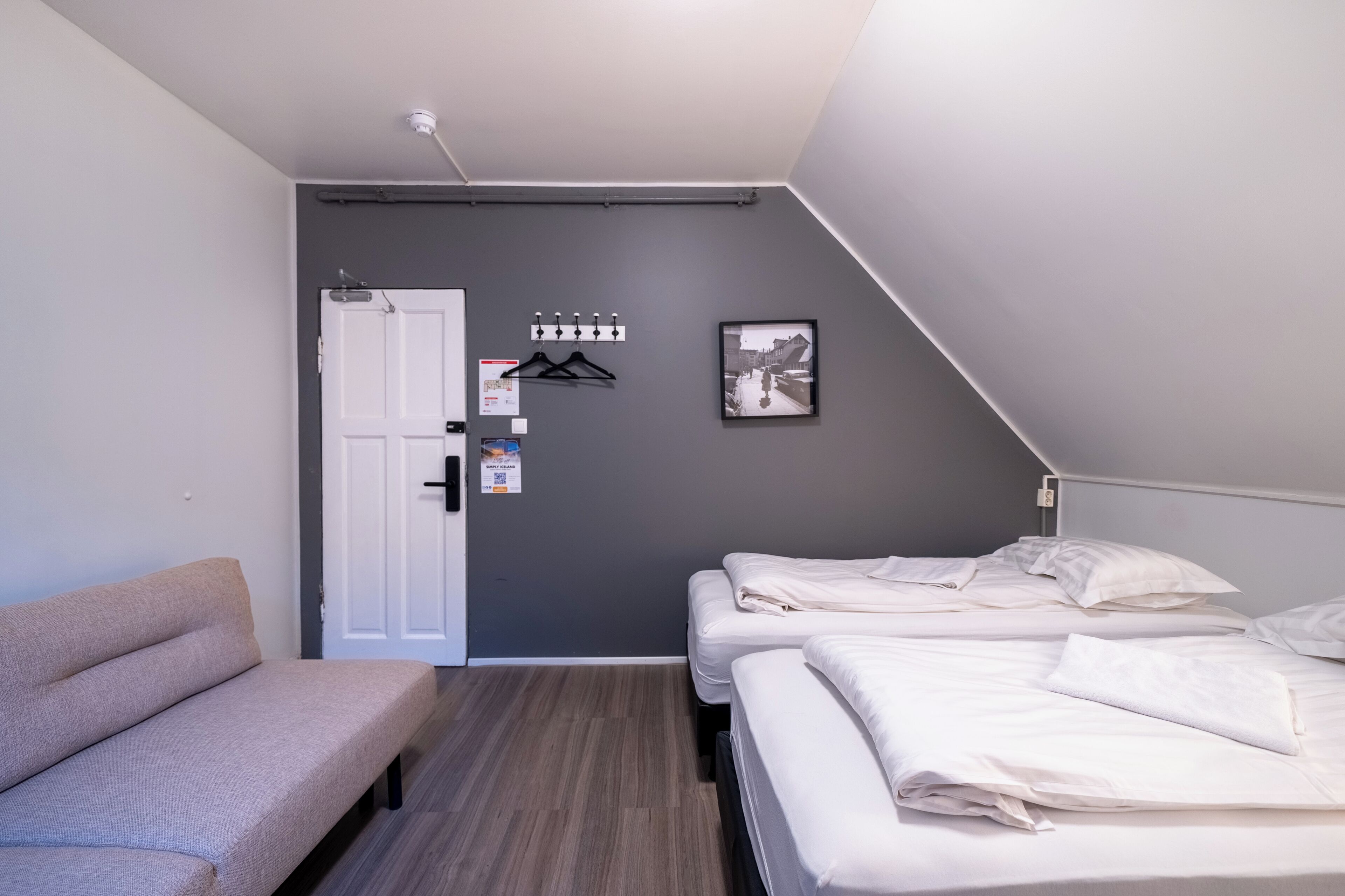 Quarto pequeno e moderno com parede cinza, porta branca, um banco e duas camas.