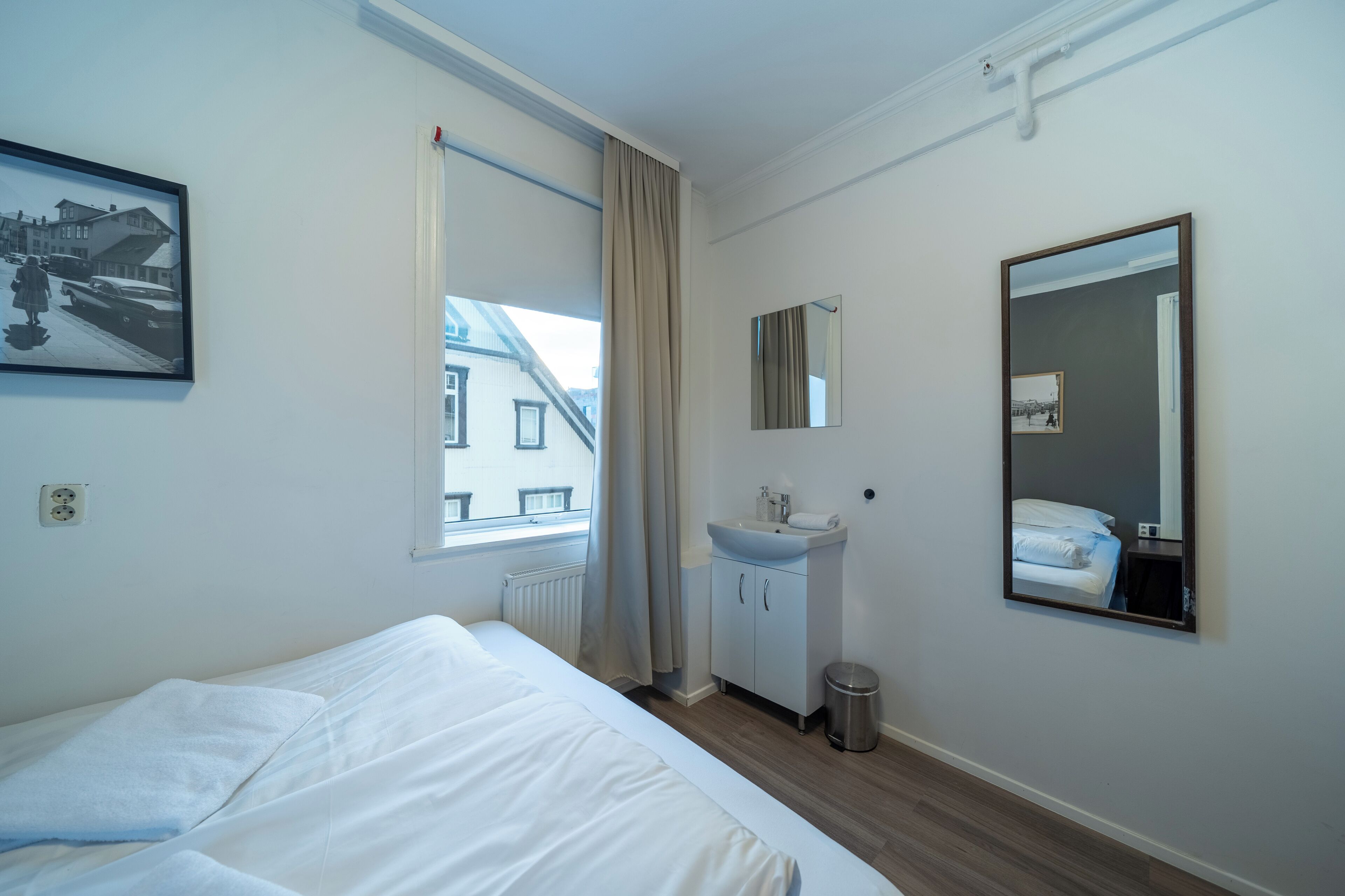 A hotel room with a bed, a window with curtains, a sink, a mirror, and a picture on the wall.