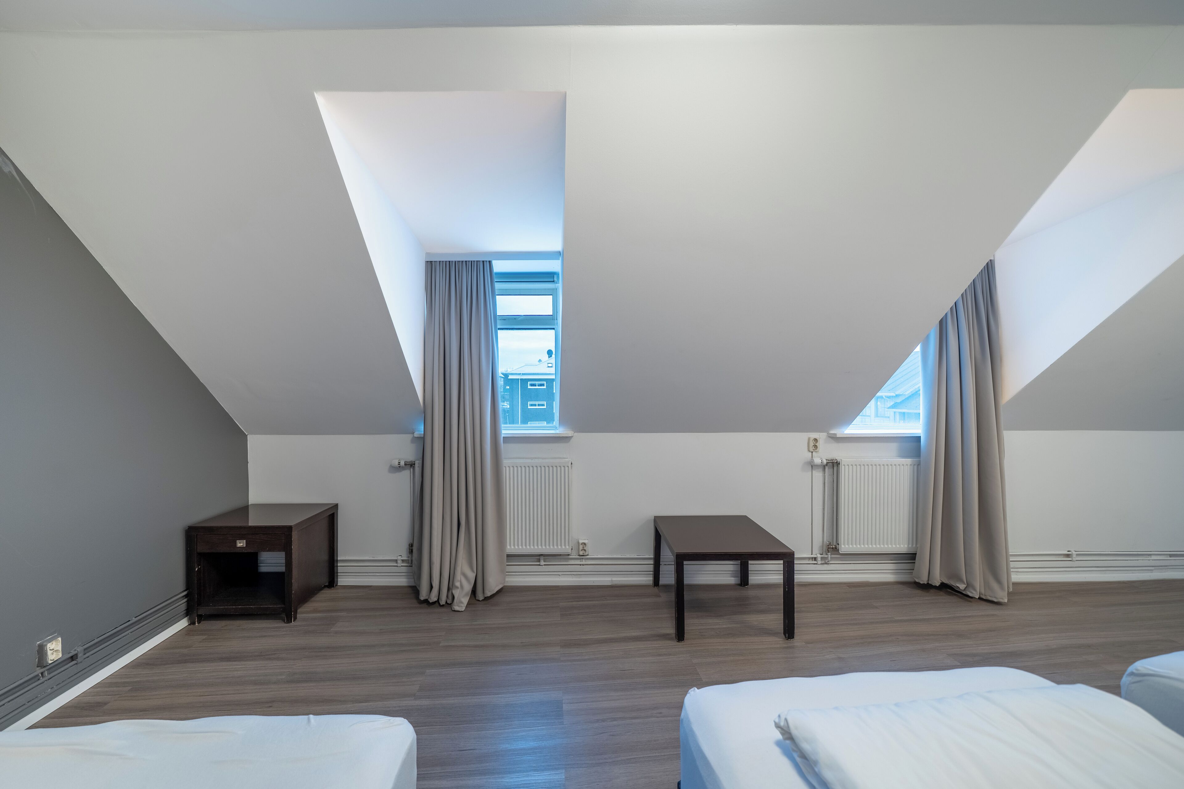 Habitación de hotel moderna con techo inclinado, dos camas, una mesita y una ventana con cortinas.