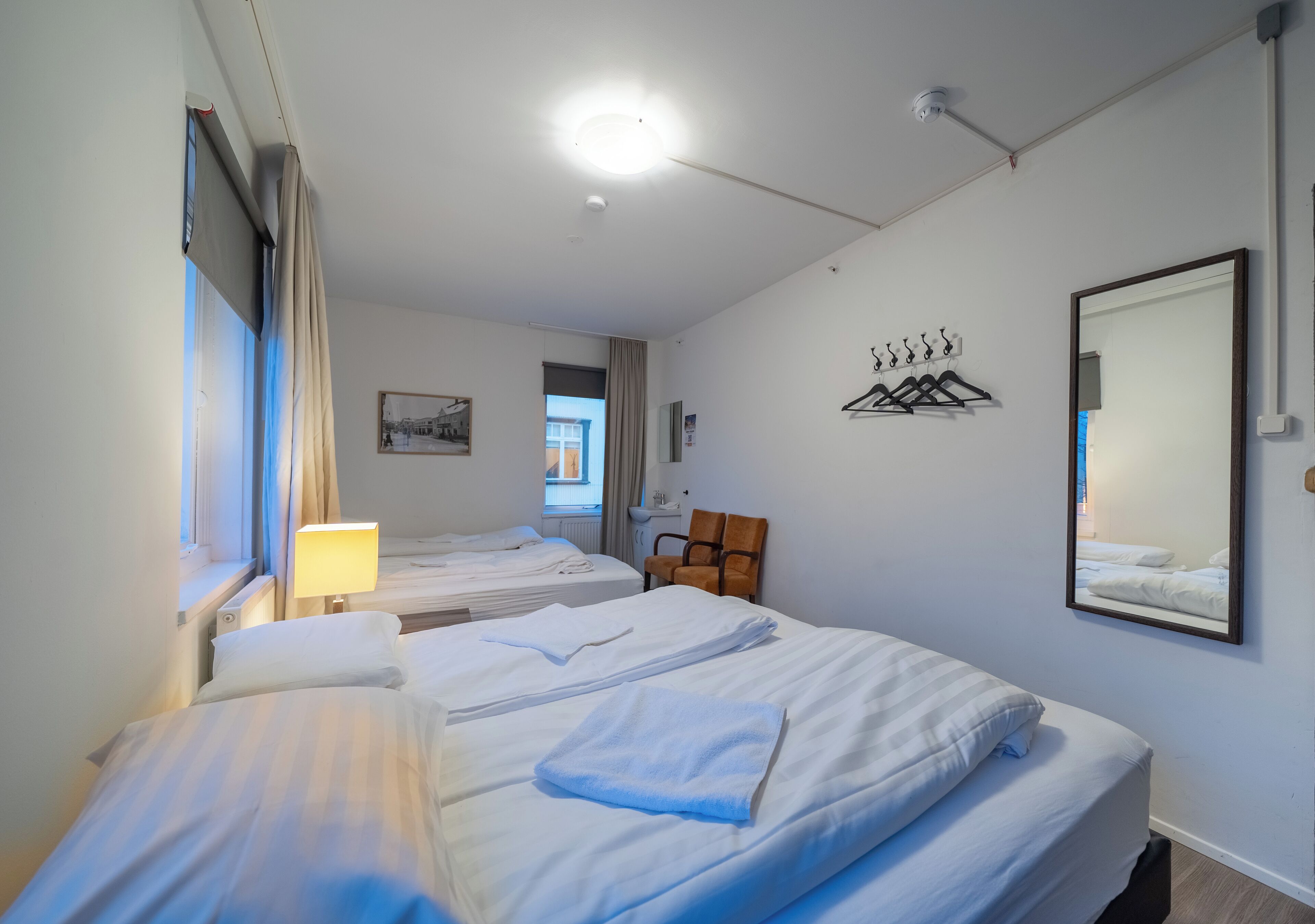 A hotel room with two beds, a chair, a mirror, and a window with curtains.