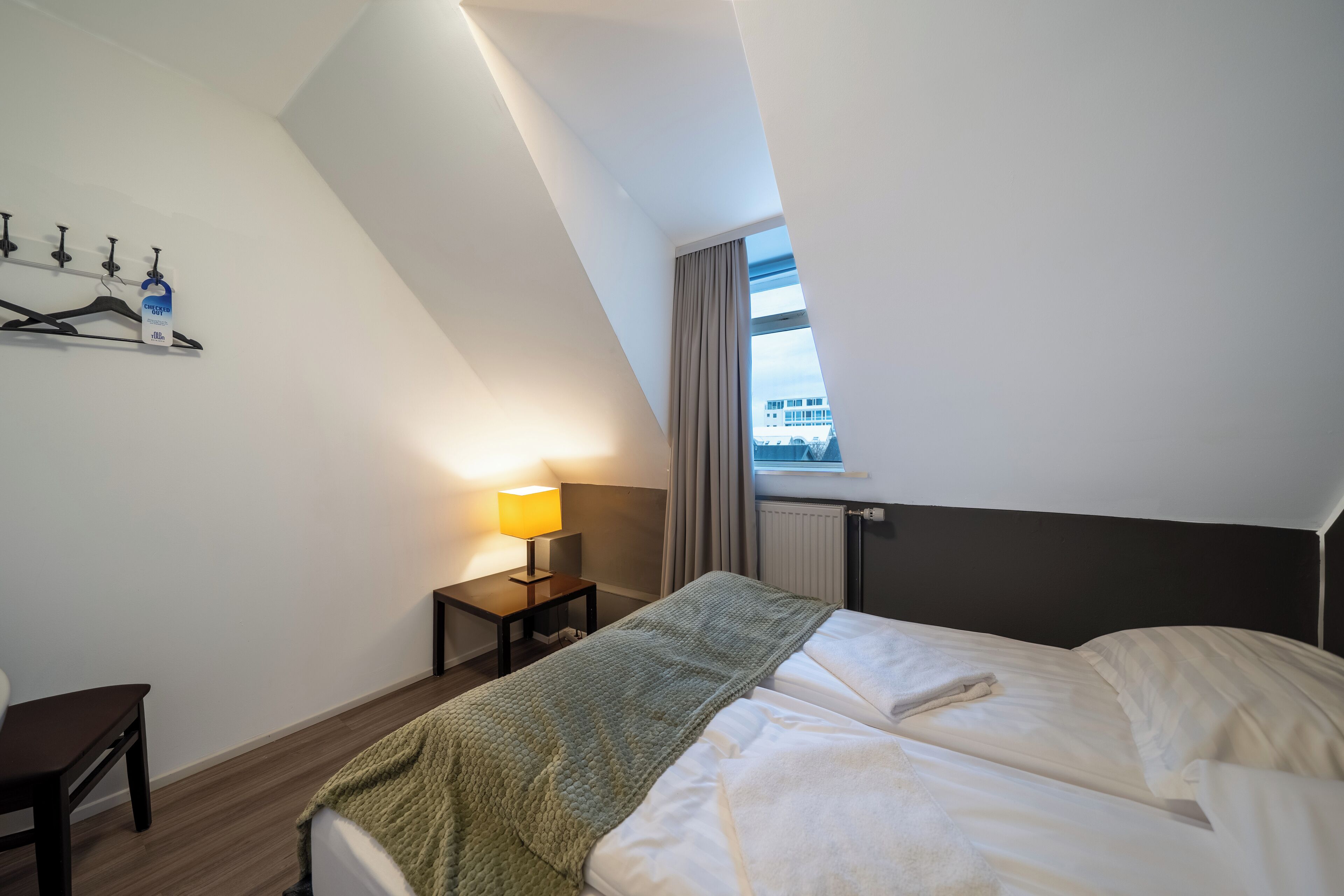 Quarto de hotel moderno com uma cama grande, uma escrivaninha, uma luminária e vista da cidade pela janela.