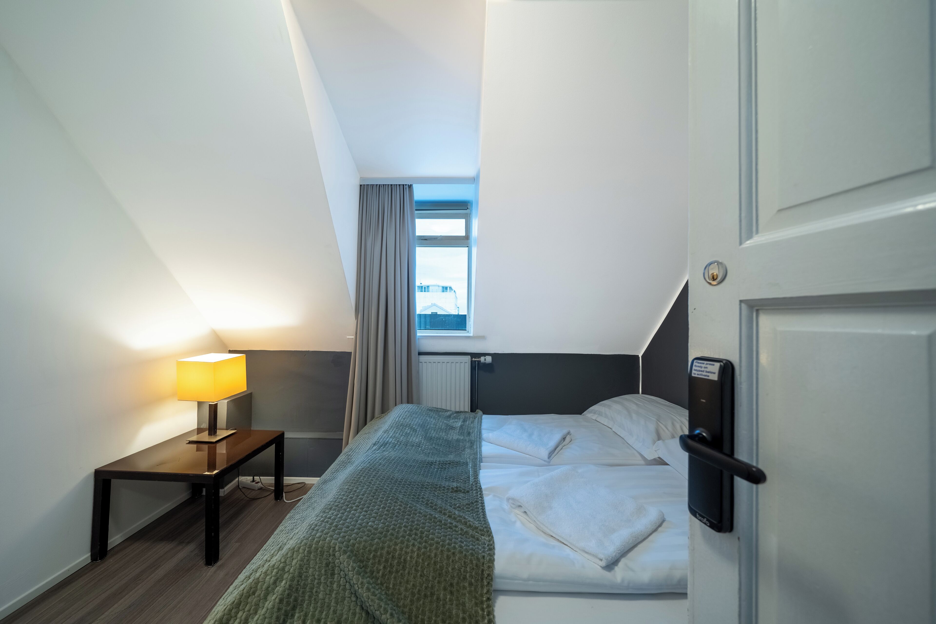 Habitación de hotel con cama, mesita de noche, lámpara y vistas al exterior por una ventana.