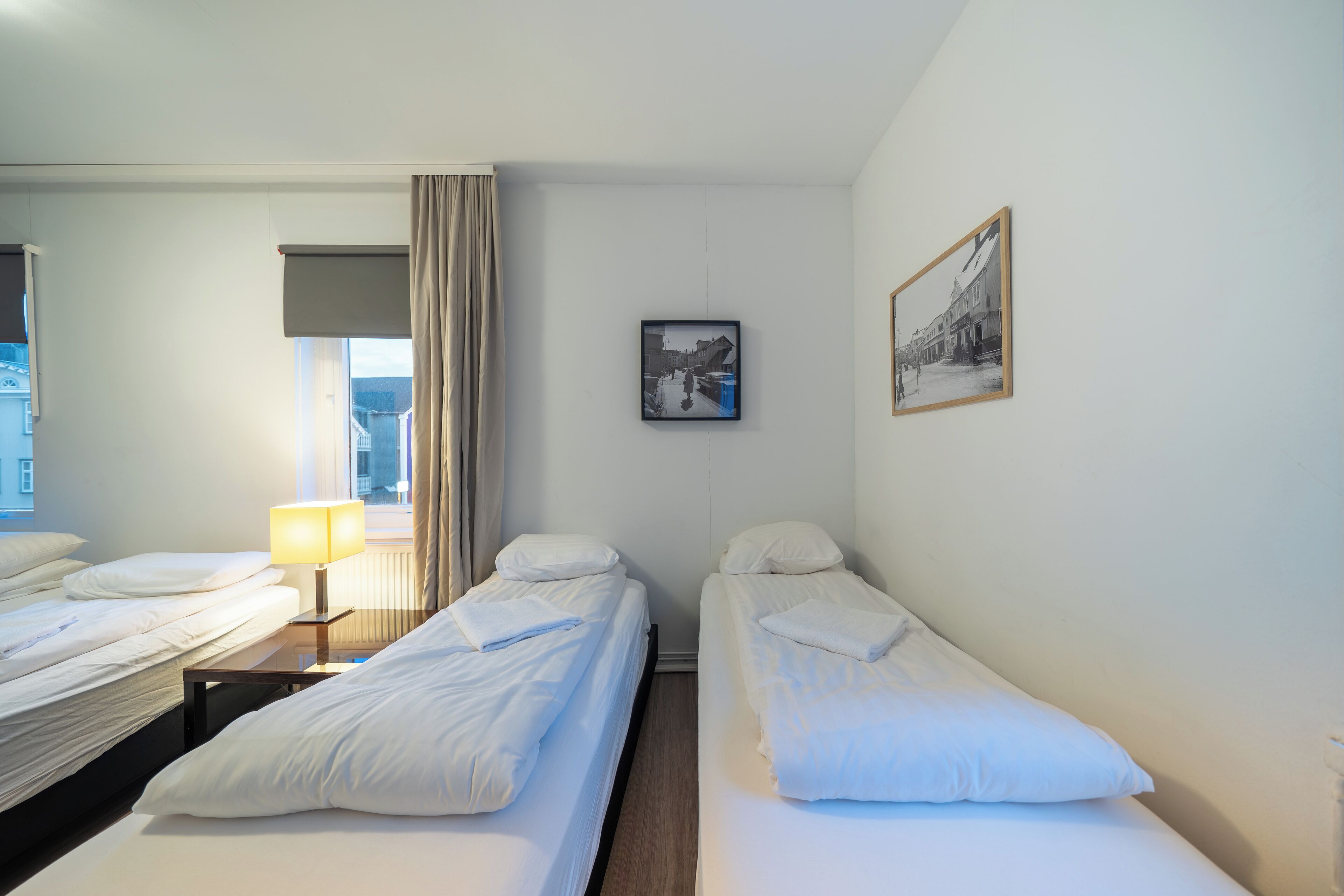 A hotel room with two single beds, a bedside table with a lamp, a window with curtains, and a framed picture on the wall.