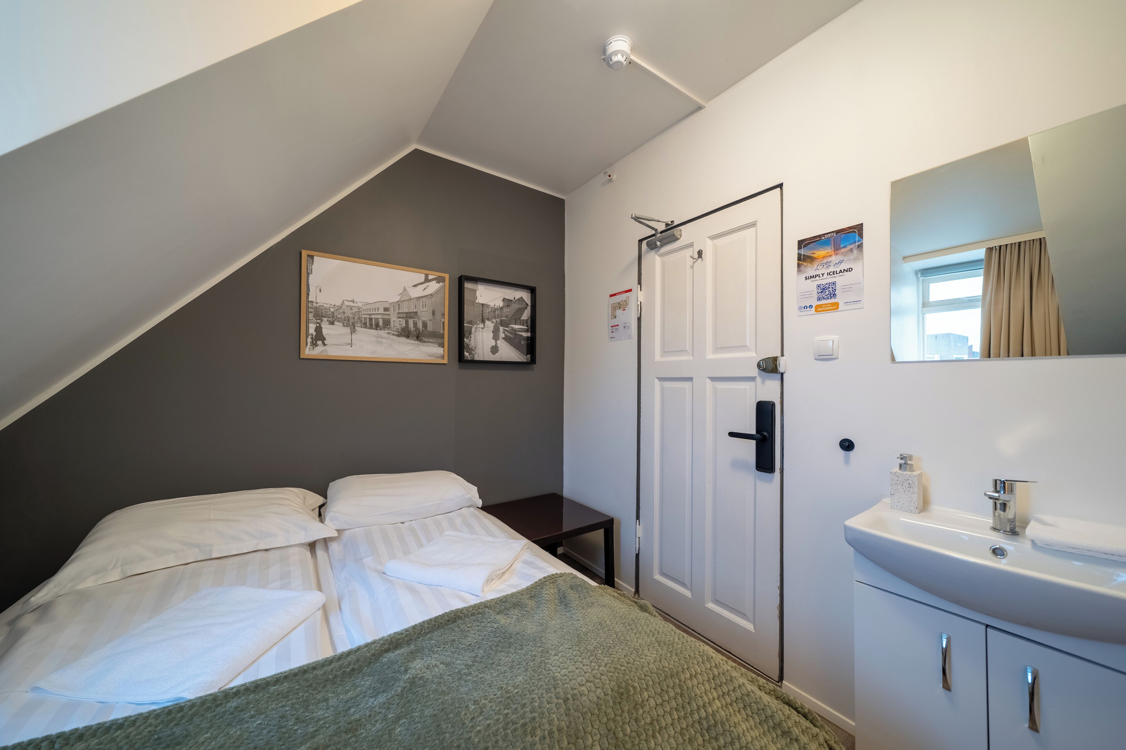 Un dormitorio con una cama, una mesita de noche, una pared blanca con una puerta, una ventana con cortinas y un cuadro enmarcado.