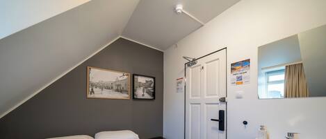 Un dormitorio con una cama, una mesita de noche, una pared blanca con una puerta, una ventana con cortinas y un cuadro enmarcado.