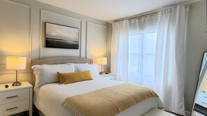 Une chambre d’hôtel bien aménagée, avec un grand lit, deux tables de chevet, une lampe de chevet, un tableau au mur et une fenêtre avec des rideaux.