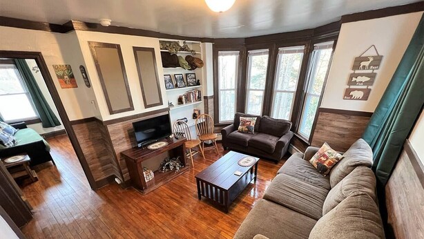 A cozy living room with a wooden floor, a sofa, a coffee table, and a TV on a wooden stand. There are windows with curtains, a bookshelf, and a wall with animal decorations. - Launch your next adventure from here: plenty of beds, plenty of parking, plenty of fun (Plymouth)