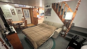 Een compacte kamer met een bed, een klein tafeltje en een stoel. Een trap met verlichting, een deur met een bordje en een muur met een schilderij.
