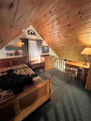 Une chambre confortable située sous les toits, avec un plafond en bois, un lit recouvert d’une courtepointe à motifs, un bureau avec une lampe et une chaise.