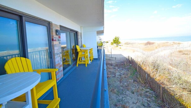 A balcony with yellow chairs and a table, overlooking a beach with a fence and sparse vegetation.