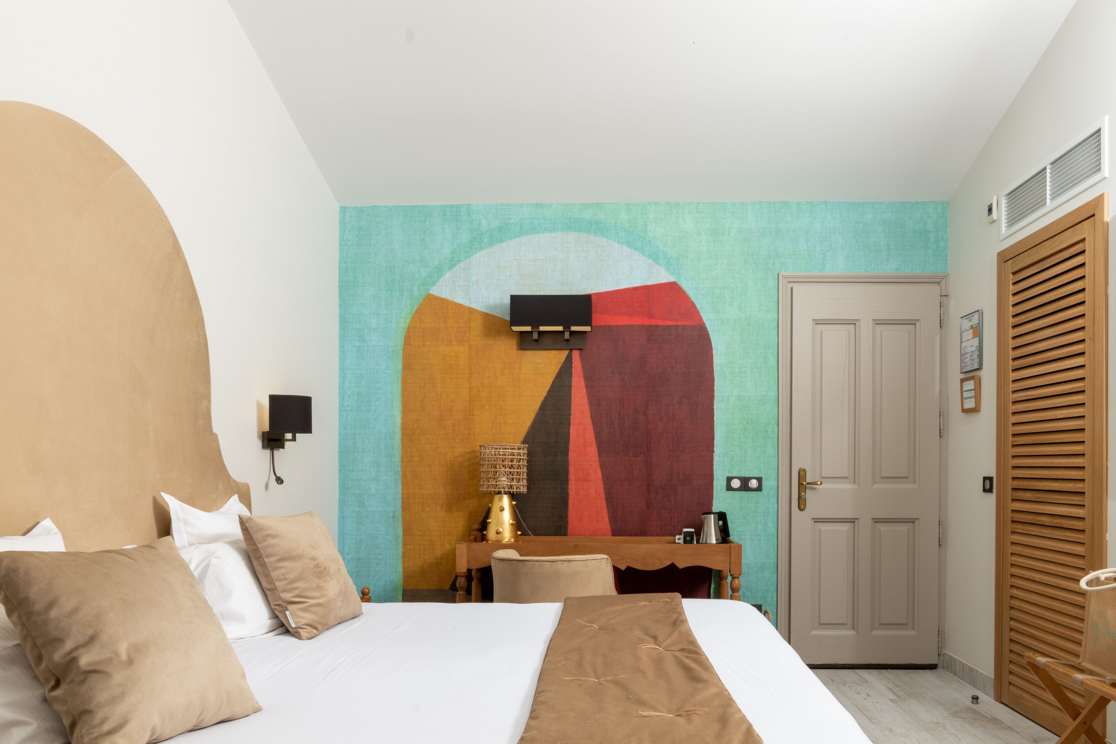 Una habitación de hotel con cama, escritorio, silla, un cuadro en la pared y una puerta.