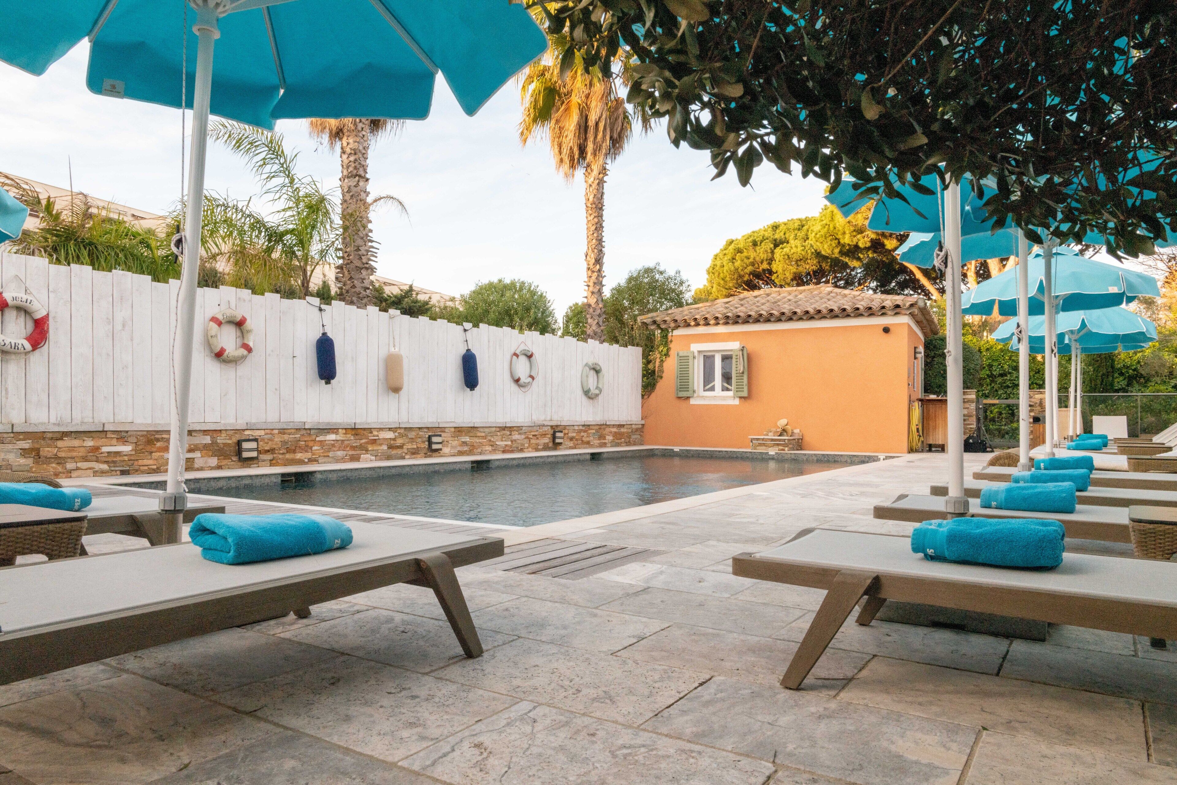 A pool area with lounge chairs and umbrellas, a white fence with life rings, and a small building with a tiled roof.
