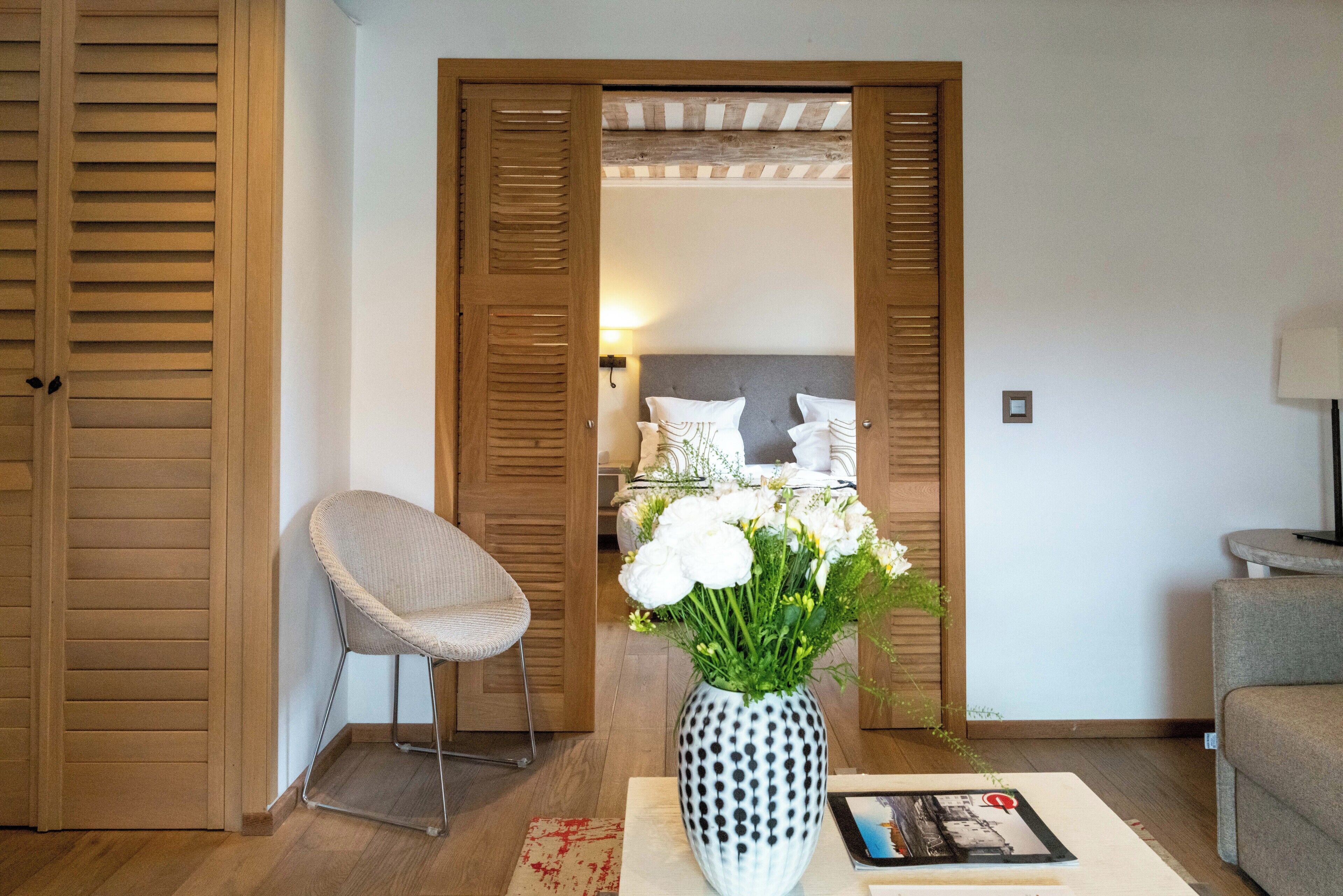 A room with a wooden door, a chair, a bedside table with a vase of flowers, and a bed with pillows and a nightstand.