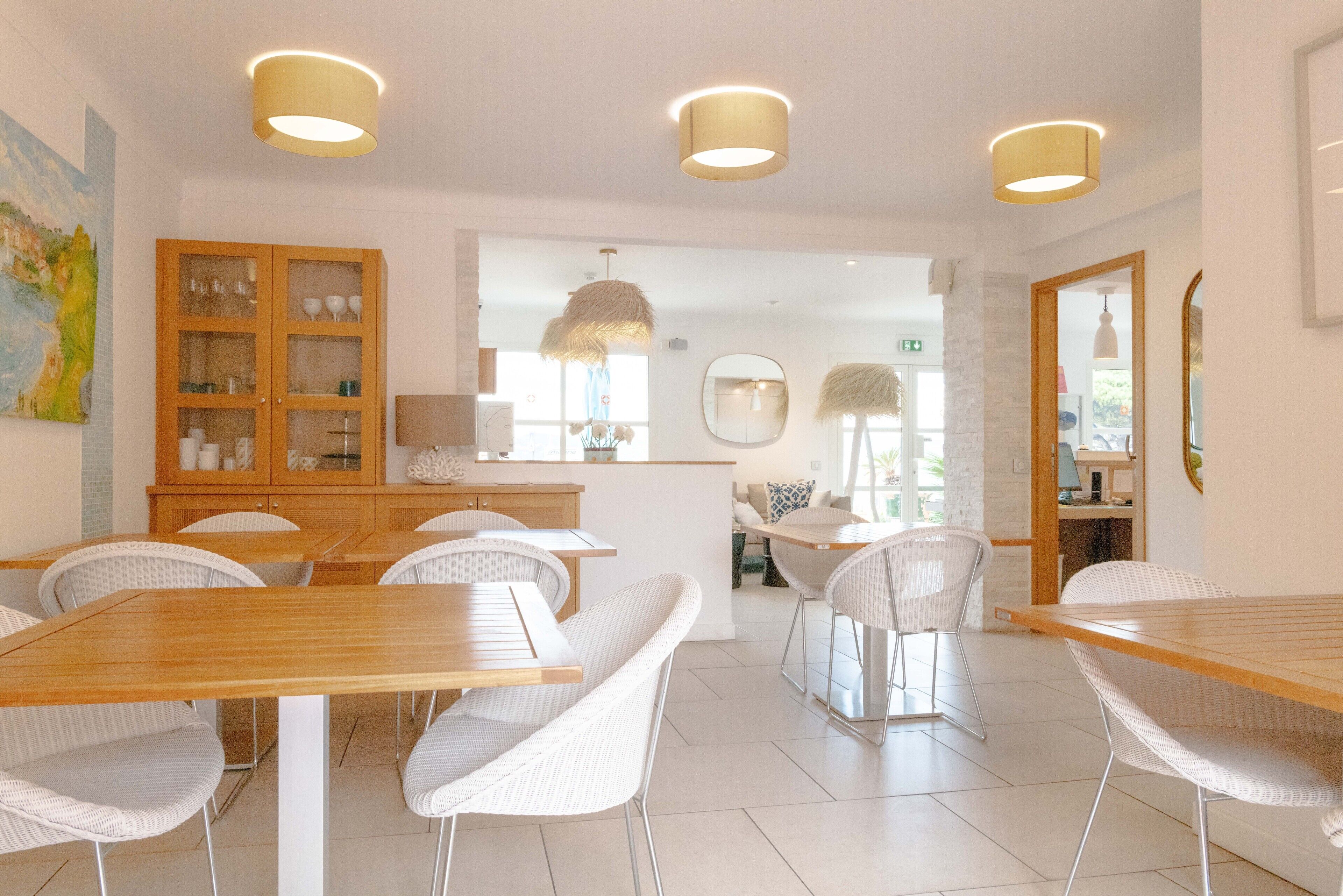 Un espace repas moderne avec des tables en bois et des chaises en osier blanc, un buffet en bois doté de portes en verre et un éclairage suspendu.
