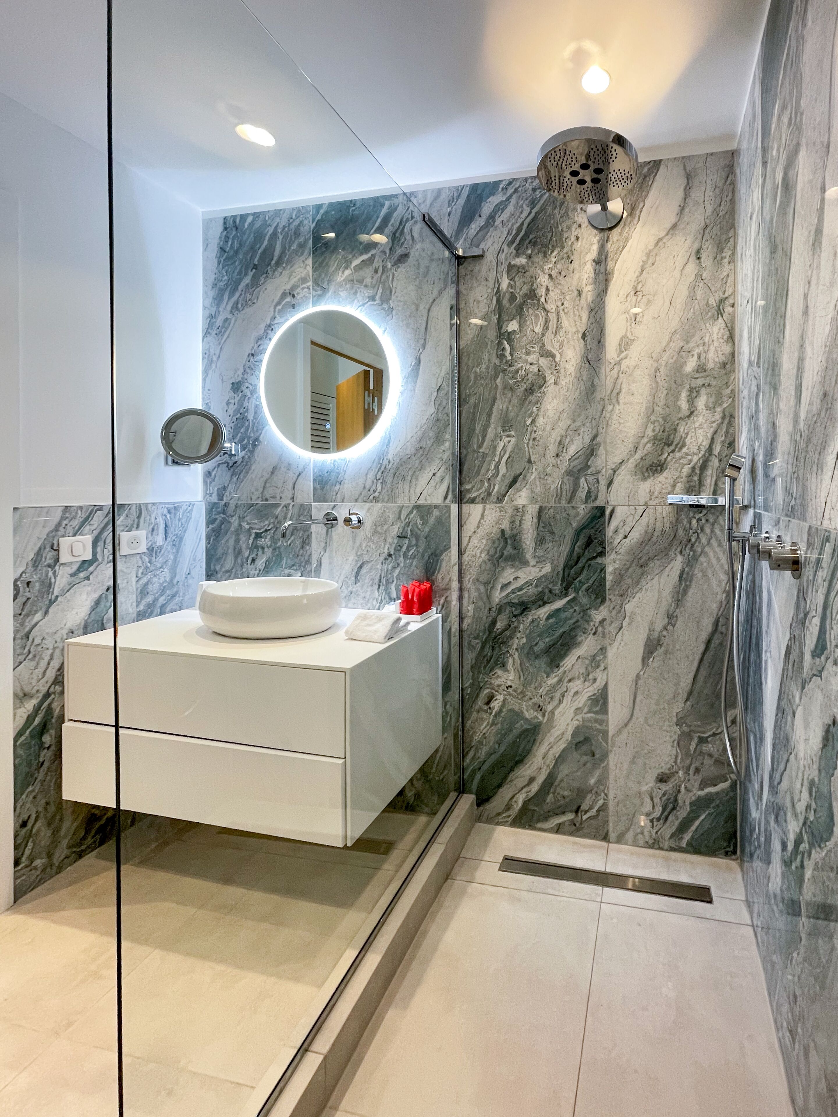 Une salle de bain moderne avec un mur en marbre, un miroir rond, un meuble-lavabo blanc et un espace douche.