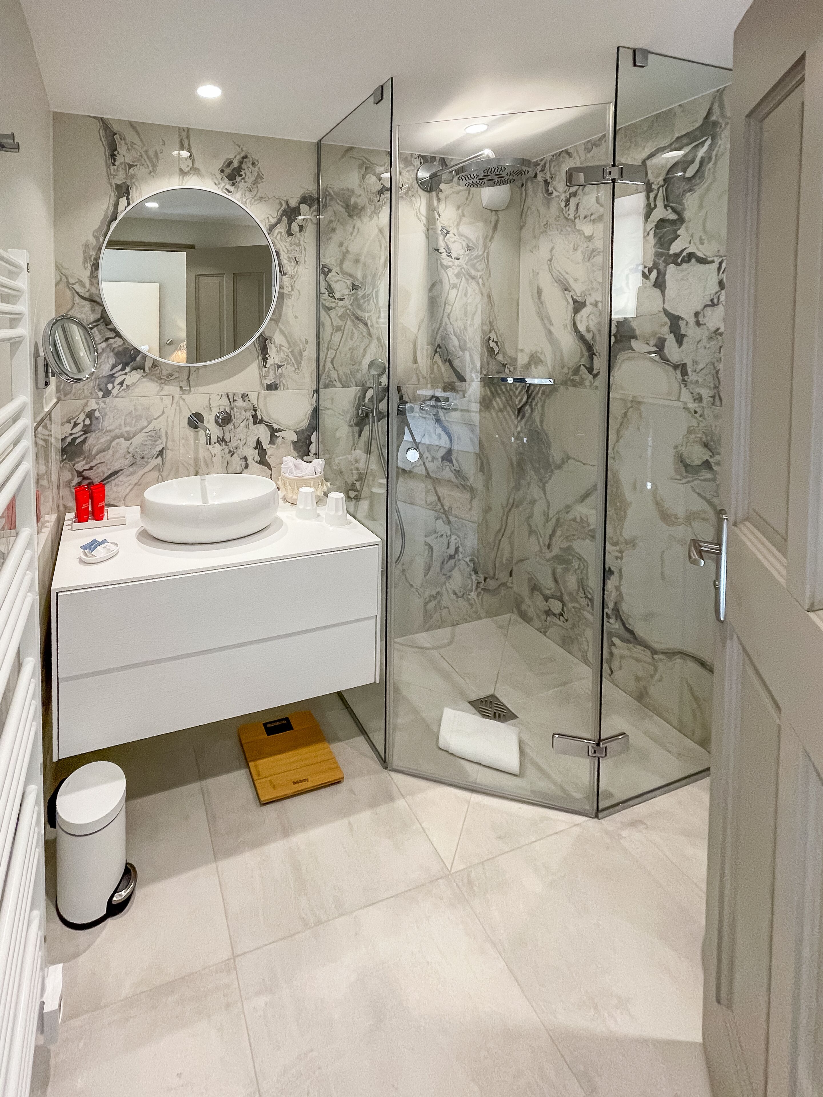 A modern bathroom with a glass-enclosed shower, a white vanity with a round mirror, and marble-patterned walls.