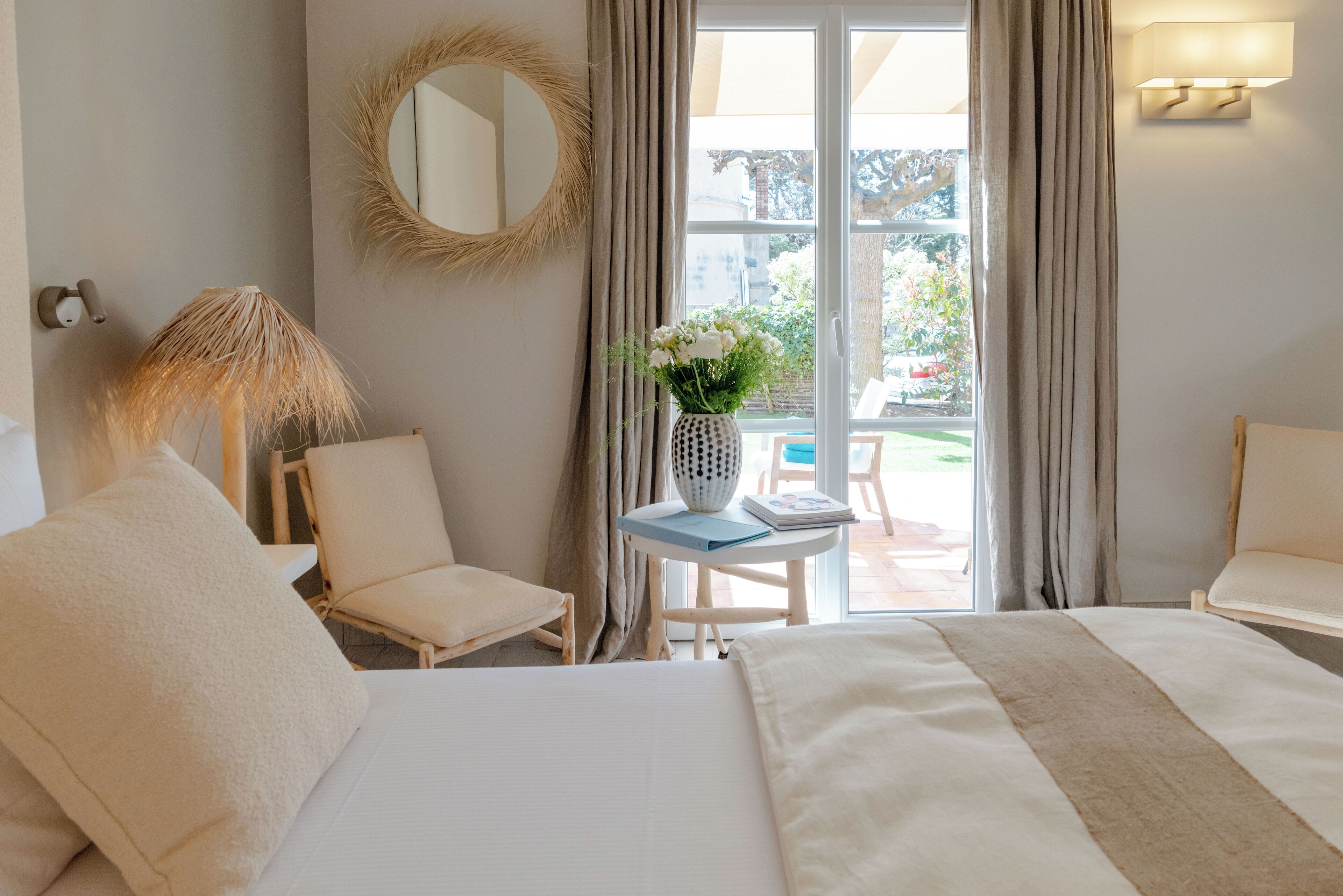 A hotel room with a bed, a chair, a small table with a vase of flowers, a round mirror, and a view of the outdoors.