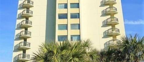 A tall, multi-story hotel with balconies on each floor, surrounded by palm trees and a well-maintained lawn.