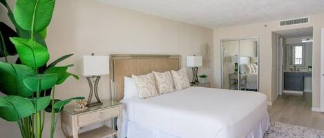 A bedroom with a large bed, bedside tables, a nightstand, a mirror, and a potted plant.