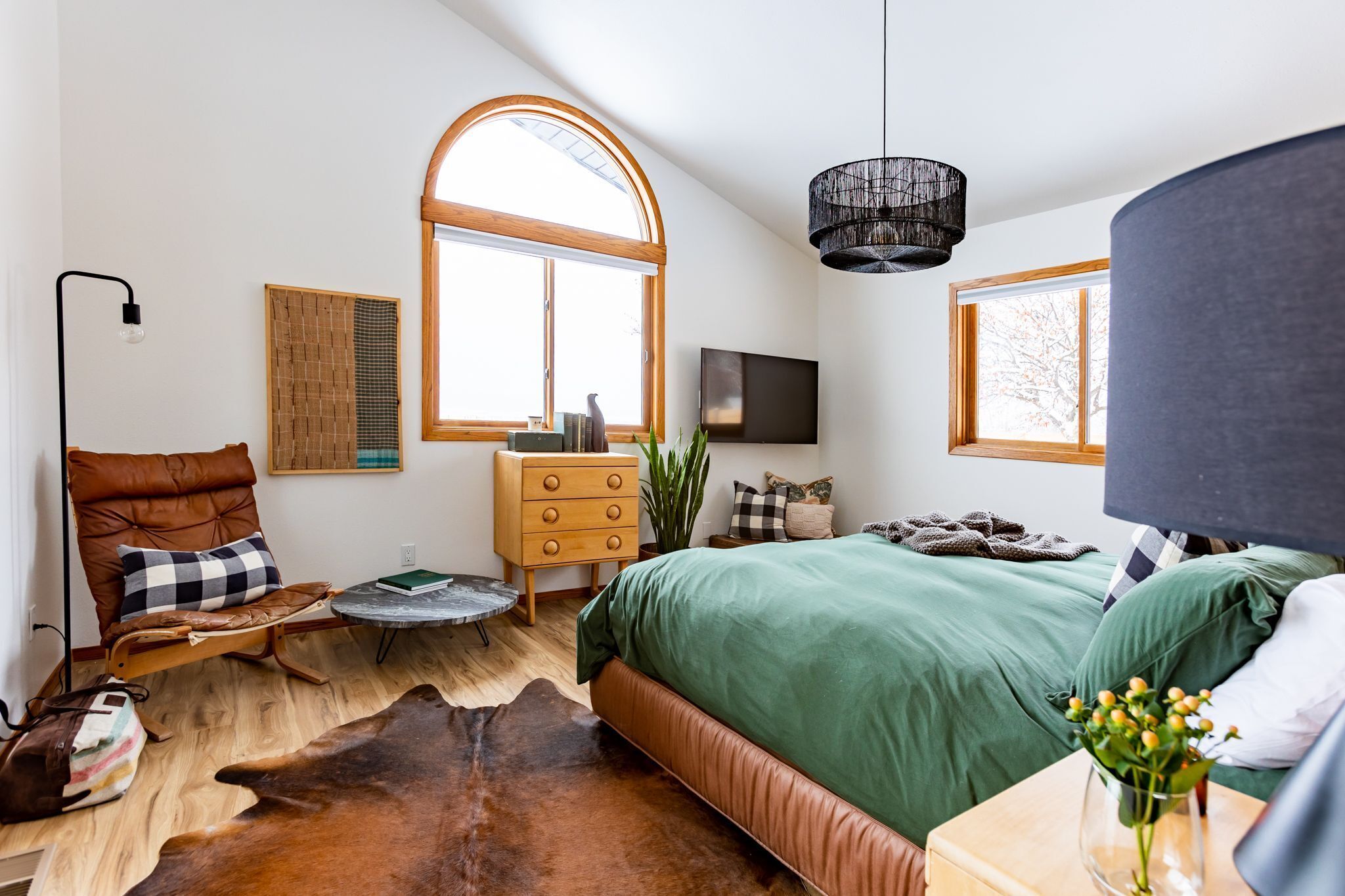 Un dormitorio moderno con una cama grande, un aparador de madera, un sillón de cuero, una mesa redonda y un ventanal amplio.
