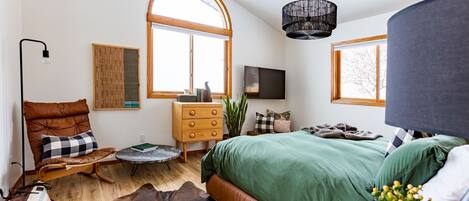 Un dormitorio moderno con una cama grande, un aparador de madera, un sillón de cuero, una mesa redonda y un ventanal amplio.