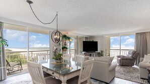 A spacious living room with a glass dining table, white chairs, a beige sofa, and a recliner. A flat-screen TV is mounted on the wall, and a large window offers a view of the ocean. - Gorgeously remodeled 2 bedroom ocean front unit - 509 South Hampton 2 Condo (Myrtle Beach)