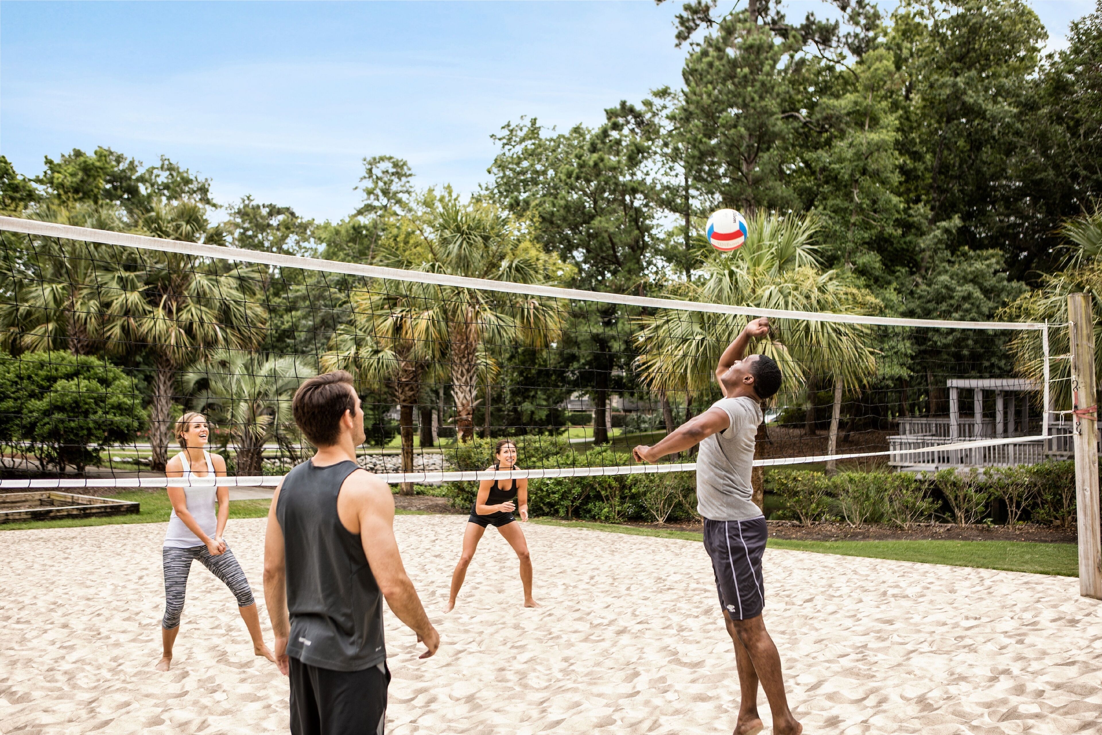 A group of people playing beach volleyball on a sandy court surrounded by trees.