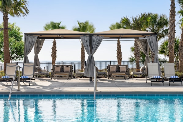 Un espace aménagé au bord de la piscine, avec des chaises longues, un auvent et des palmiers.