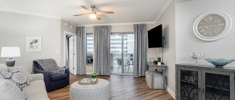 A modern living room with a ceiling fan, a round ottoman, a grey sofa, a TV, and a cabinet with decorative items.