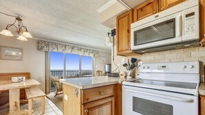 A kitchen with wooden cabinets, a microwave, an oven, and a dining area with a bench and table. - Bimini 107 2 Bedroom Condo by RedAwning (Ocean City)