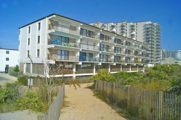 A multi-story hotel with balconies, a sandy pathway, and a wooden fence. - Bimini 107 2 Bedroom Condo by RedAwning (Ocean City)