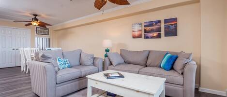 A living room with two grey sofas, a white coffee table, and a ceiling fan. There are three wall-mounted paintings and a lamp on a side table.