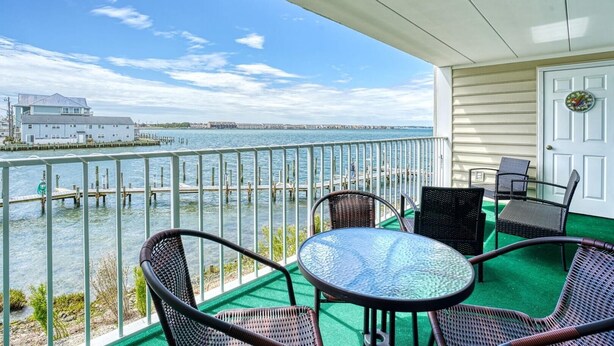 A balcony with wicker chairs and a table, overlooking a waterfront with docks and a white building in the distance.