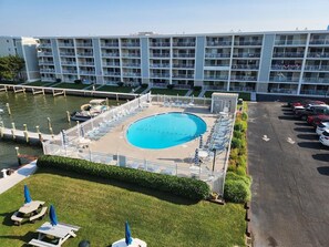 Piscina com espreguiçadeiras e guarda-sóis, cercada por uma grade, com um estacionamento e um edifício ao fundo.