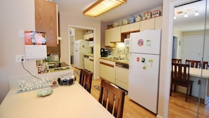 A compact kitchen with wooden cabinets, a refrigerator, a microwave, and a dining area with a table and chairs. - Club Ocean Villas Ii 19 2 Bedroom Condo by RedAwning (Ocean City)