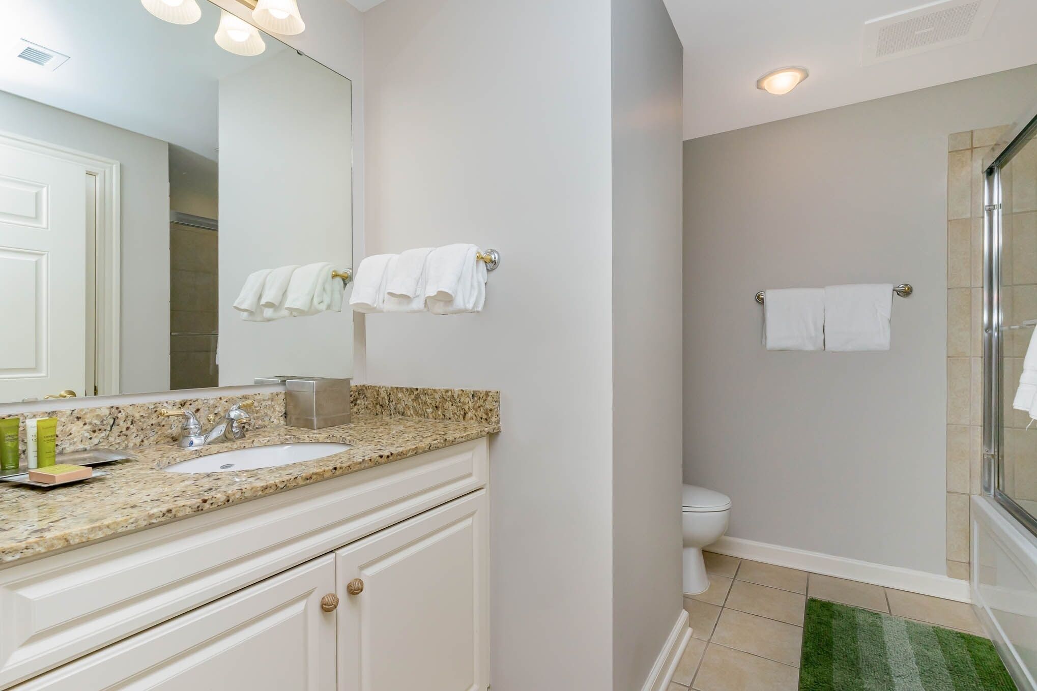 A bathroom with a granite countertop sink, white cabinets, a mirror, and a shower area.