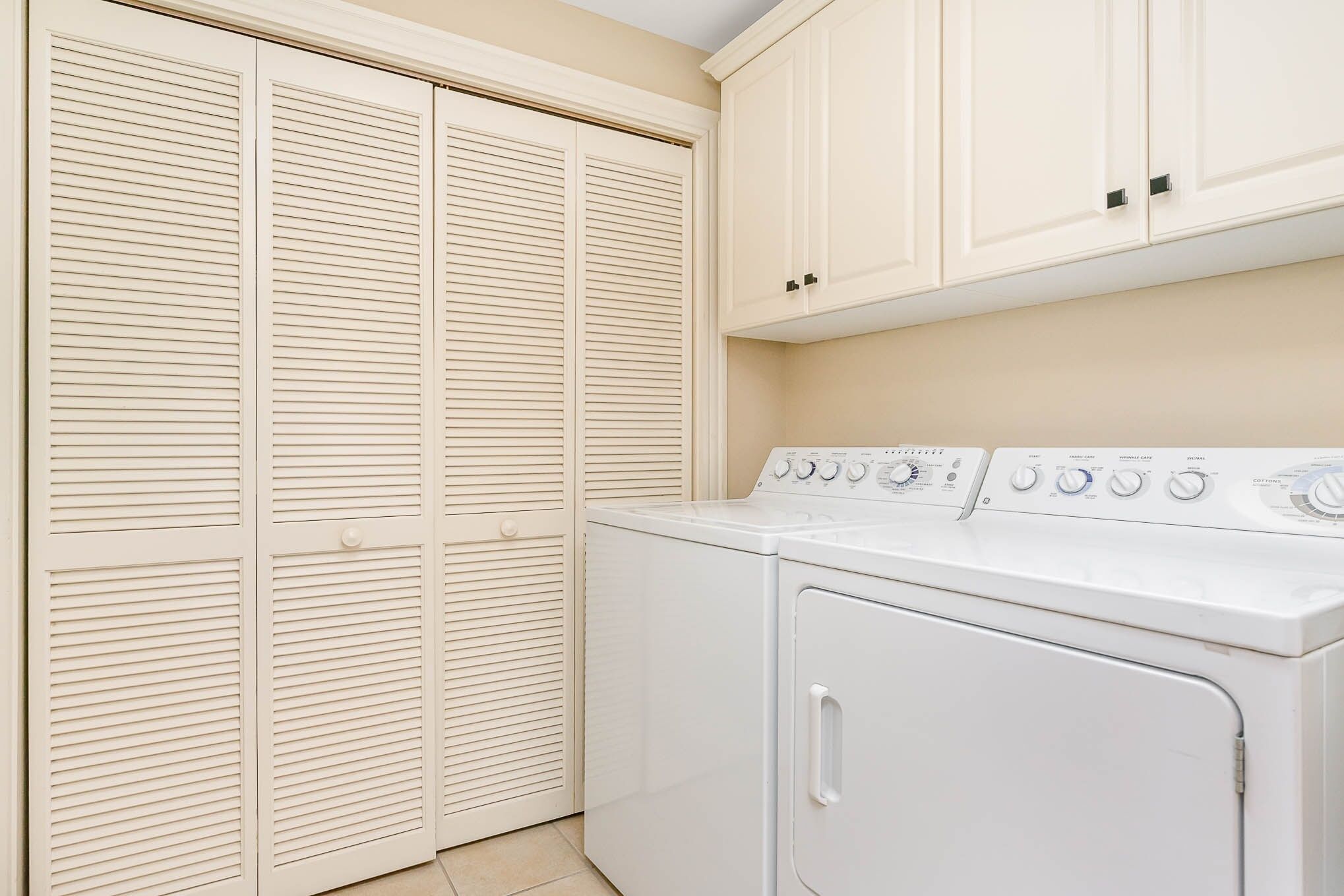 A laundry room with a front-loading washer and dryer, beige walls, and a white door with horizontal louvered panels.