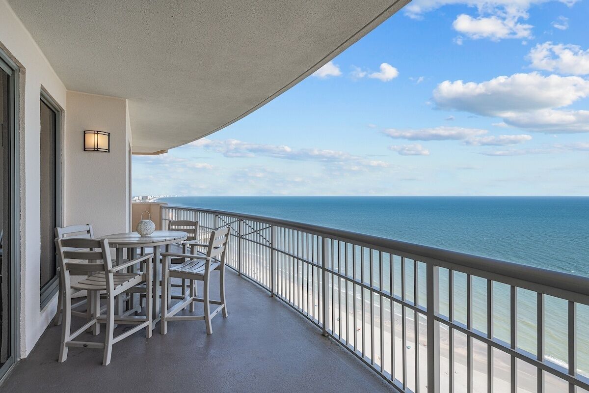 A balcony with a table and chairs, overlooking a beach and ocean.