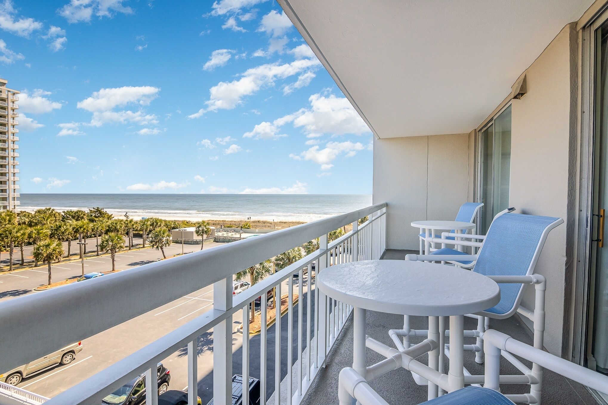 A balcony with white chairs and a table, overlooking a beach and ocean.