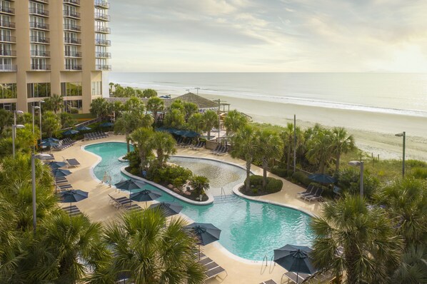Relax by the oceanfront pool surrounded by palm trees and lounge chairs, with stunning views of the ocean from the resort's multi-story building.