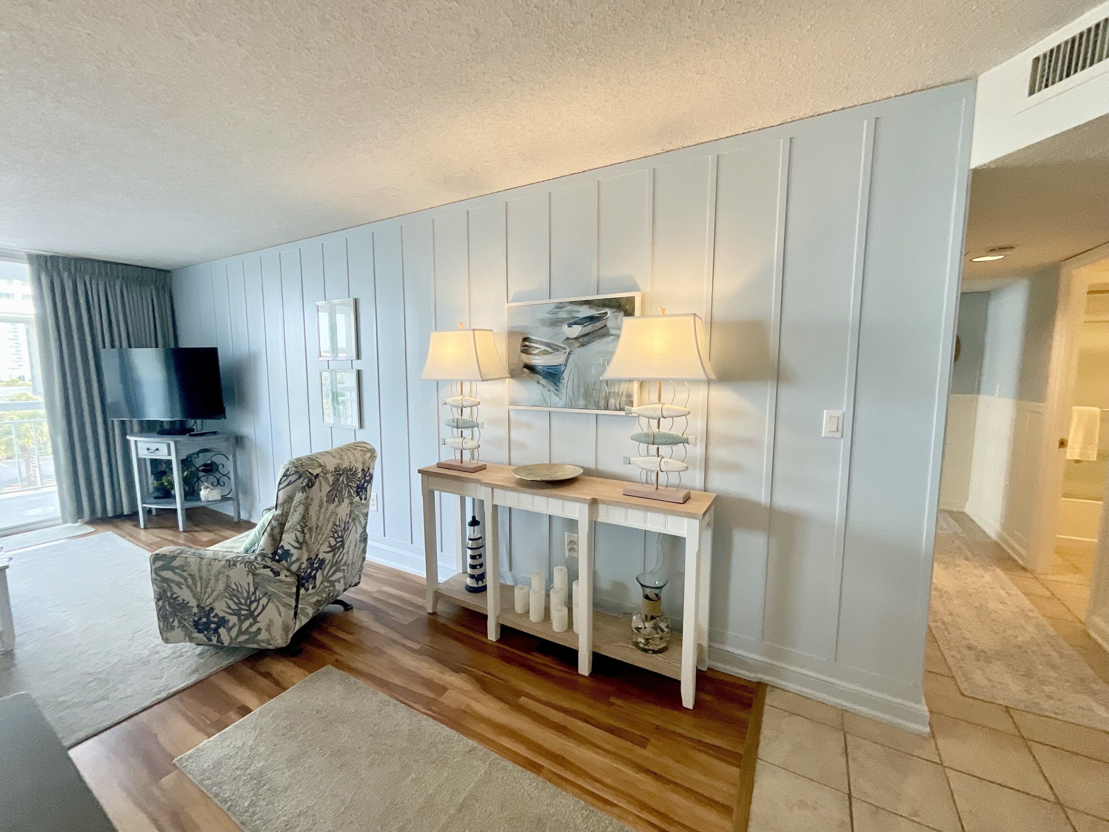 A living room with a TV, patterned armchair, console table with lamps, and a painting.