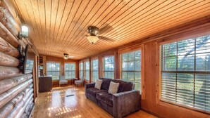 A log cabin interior with a wooden ceiling, large windows, a sofa, and a fireplace.