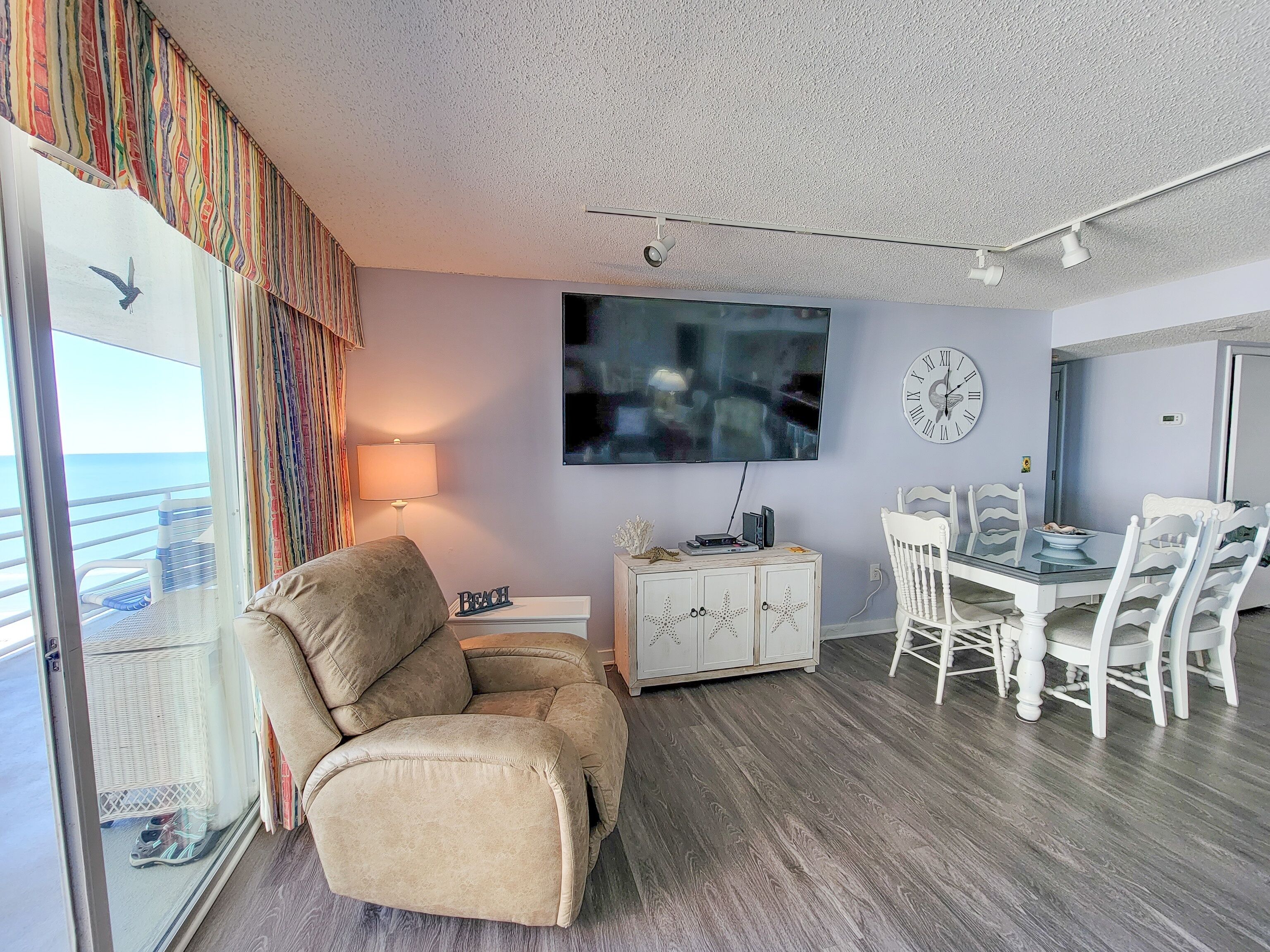 A living room with a beige armchair, a dining table with white chairs, a flat-screen TV, and a wall clock.