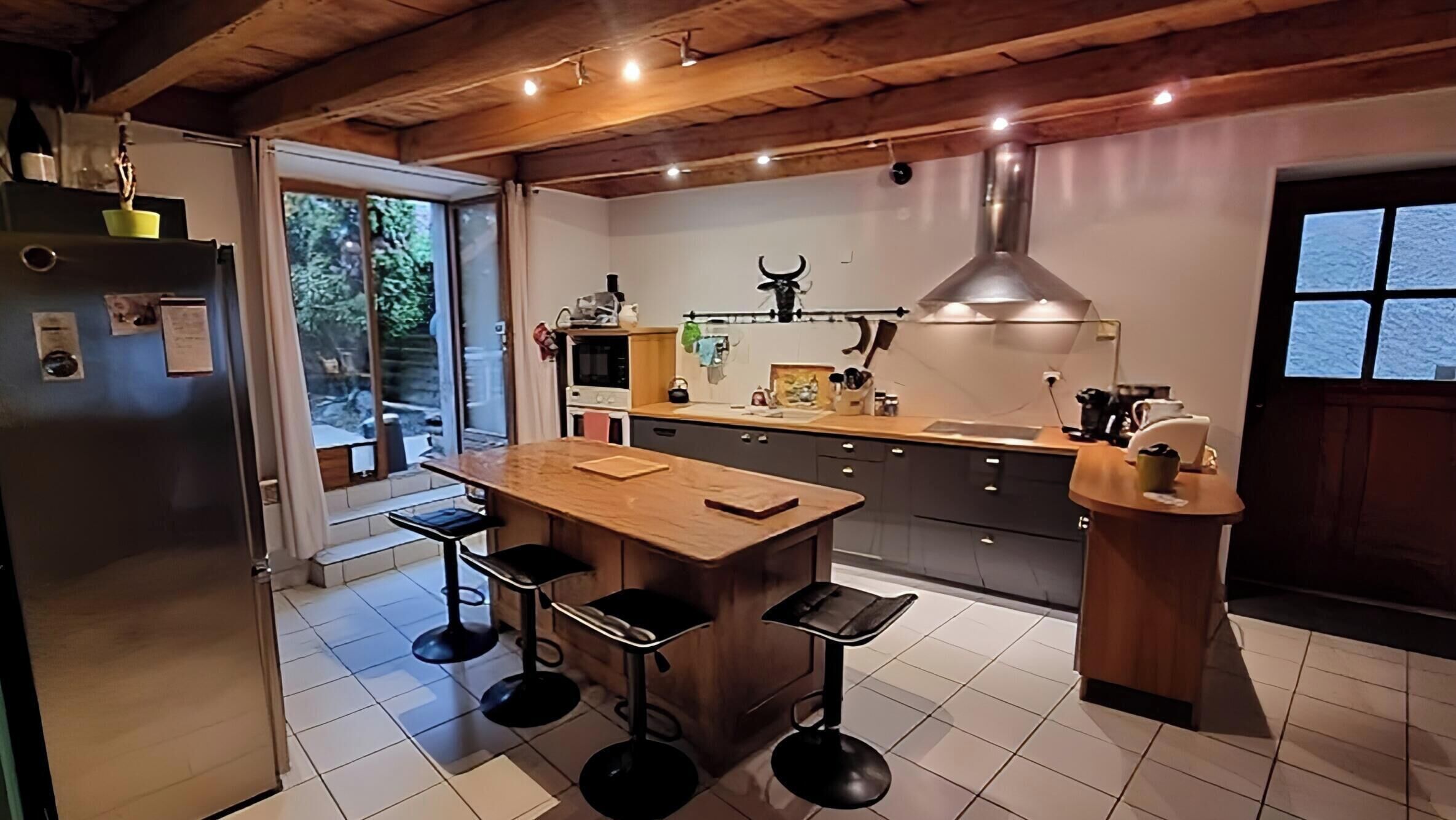 A modern kitchen with a wooden island, bar stools, a large window, and a view of the outdoors.